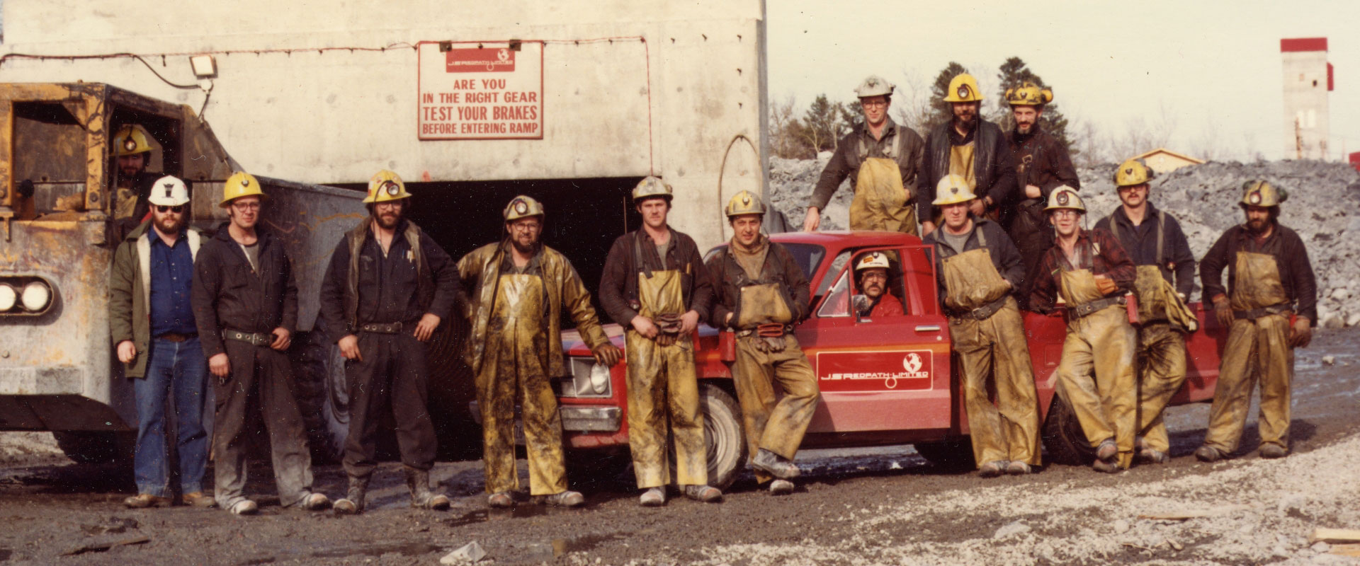 Un grupo de mineros, todos usando cascos duros con linternas, y la mayoría llevando monos o trajes de trabajo, posan orgullosamente junto a una camioneta de la compañía en frente de una gran entrada de la mina.