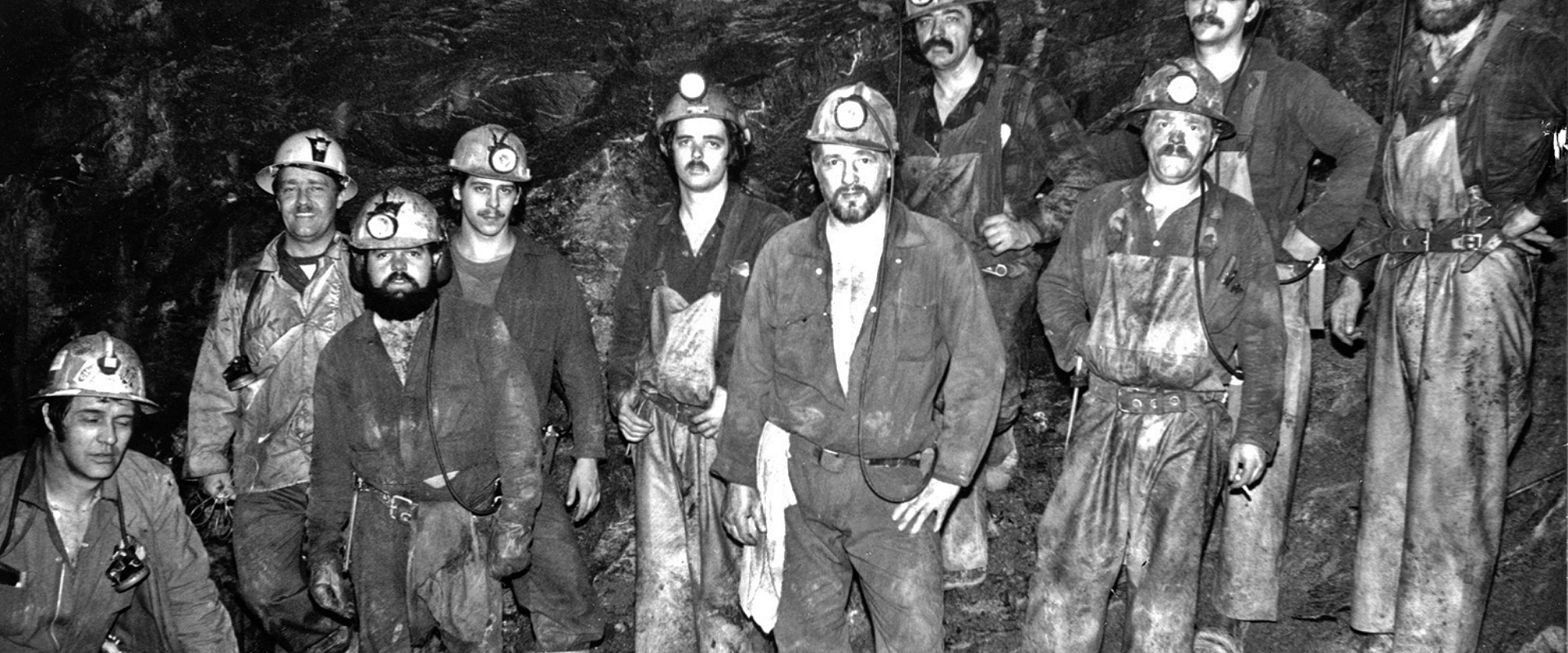 A black-and-white photograph of a group of miners of a diverse range of ages and appearances, all clad in similar work attire: overalls, work shirts, and hard hats with attached lamps, posing closely together for the camera in an underground mine, exuding a sense of unity and shared experience.