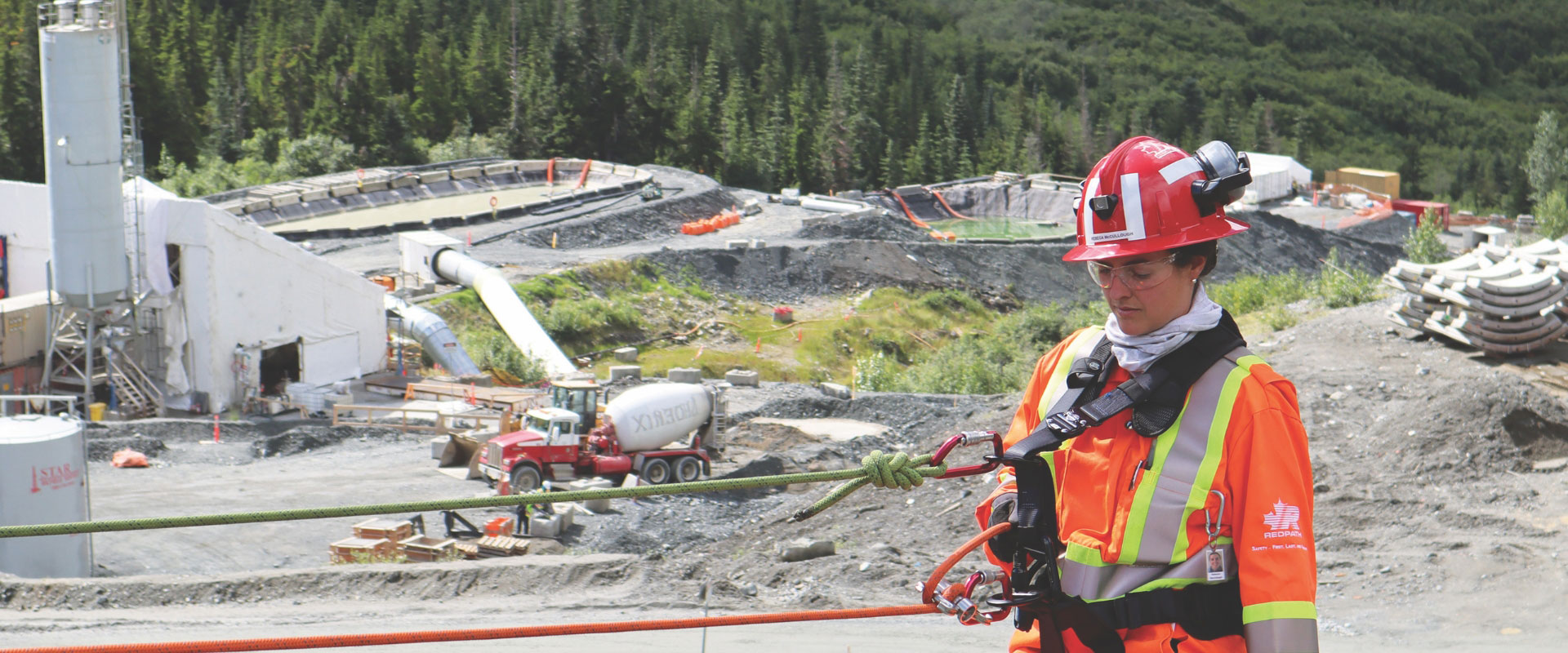 A Redpath employee is standing outside of a mine site, dressed heavily in safety gear, inspecting the equipment attached to her.