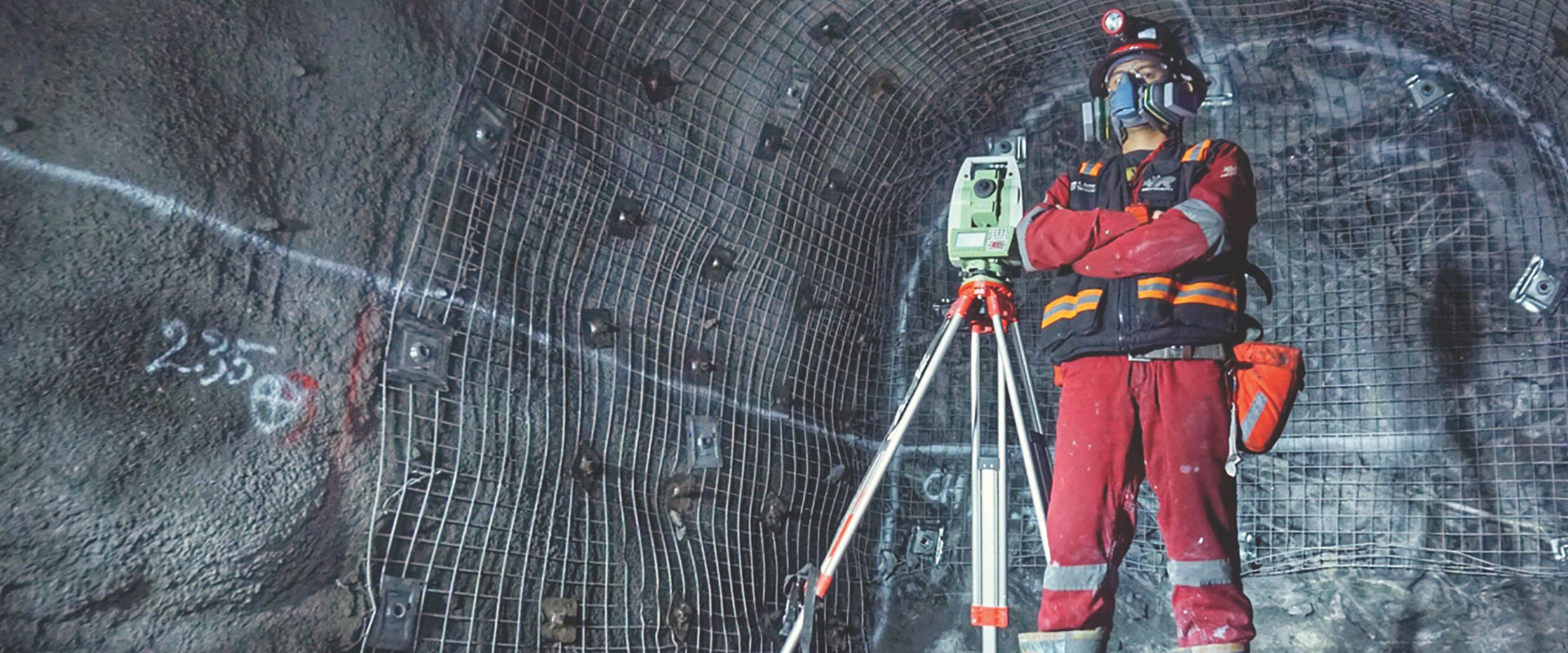Un empleado de Redpath, vestido fuertemente con equipo de seguridad, incluyendo un casco, gafas y una máscara de respiración, utiliza equipos de topografía para mapear ángulos y distancias en un túnel subterráneo.
