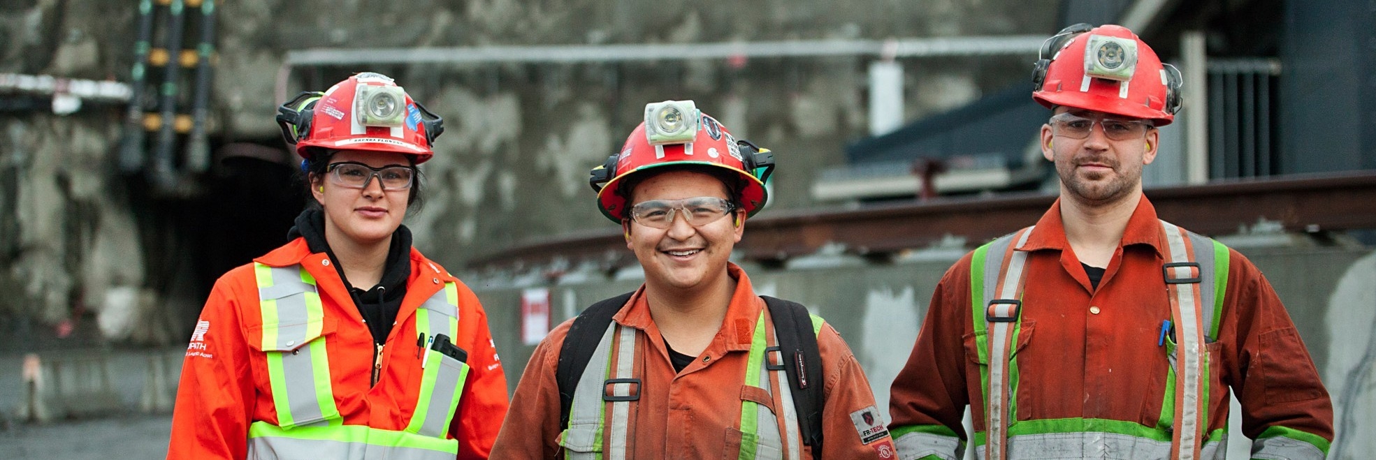 Tres empleados indígenas de Redpath parados frente a un lugar de trabajo, vistiendo gafas de seguridad y cascos protectores, sonriendo y mirando con optimismo y confianza a la cámara.
