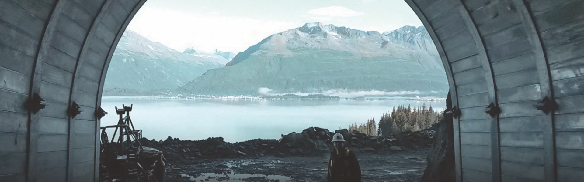 An image of a Redpath employee standing in the foreground of the opening of a mine shaft, with a natural landscape in the background, which includes mountains, forests and a lake.
