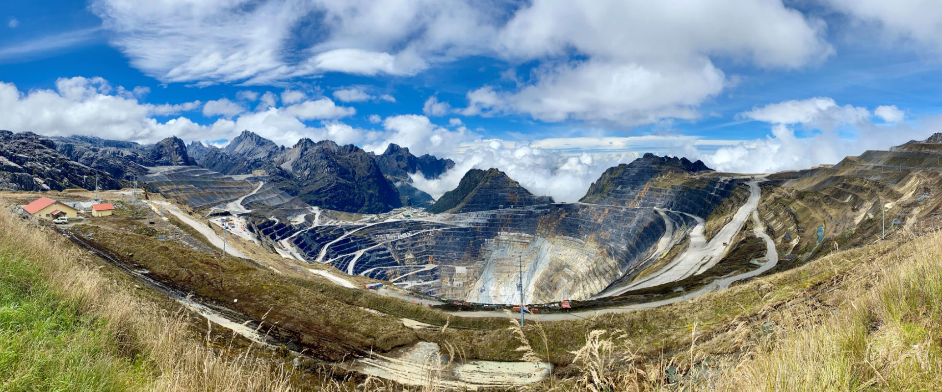 Une photo panoramique d’une énorme mine à ciel ouvert indonésienne avec des routes en terrasses superposées pour l’accès des véhicules, entourée de montagnes et de nuages.