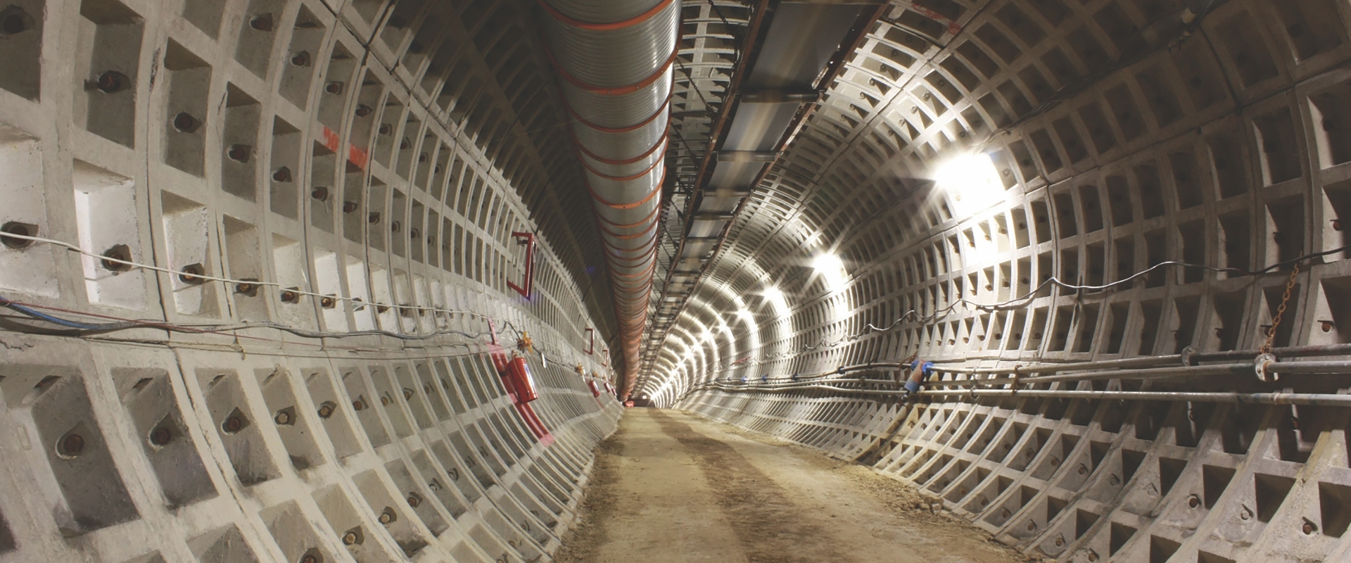 Un tunnel souterrain o&ugrave; les murs sont construits &agrave; partir d&rsquo;un motif de rectangles encastr&eacute;s, dispos&eacute;s en grille qui s&rsquo;incurvent le long de l&rsquo;arc du tunnel. Le long du sommet du tunnel, un grand tuyau ondul&eacute; court sur toute la longueur de la vue, soutenu par une charpente de poutres m&eacute;talliques et de fils.